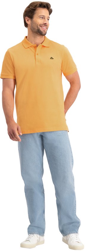 Man met oranje poloshirt, lichtblauwe jeans en witte sneakers, met handen achter de rug.