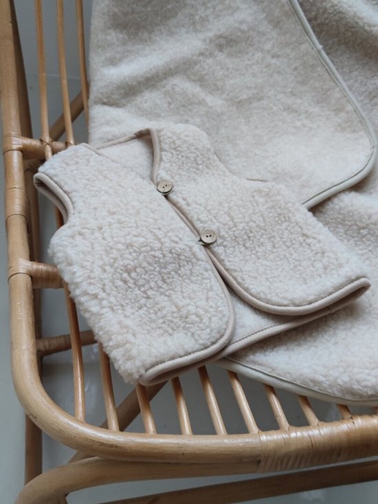 Crèmekleurig wollen kinder vest met bruine houten knopen, op een rieten stoel.