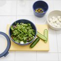 Bol Mepal bleu avec salade, lamelles de concombre, pistaches et petites billes de mozzarella sur planche à découper jaune.