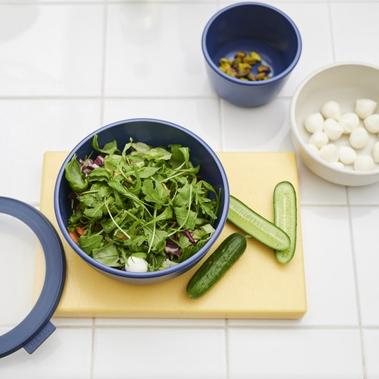 Bol Mepal bleu avec salade, lamelles de concombre, pistaches et petites billes de mozzarella sur planche à découper jaune.