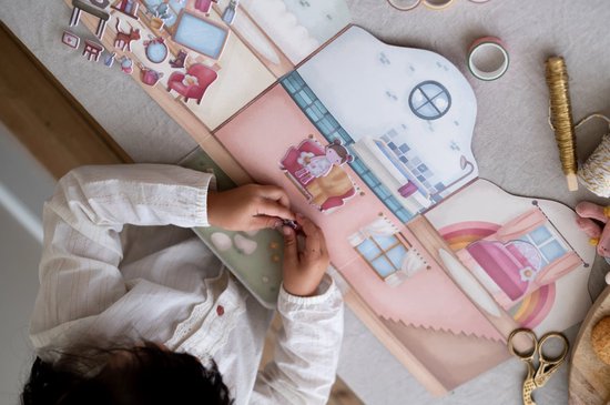 Meisje plakt stickers in een kartonnen poppenhuis speelset met kamers en meubels.