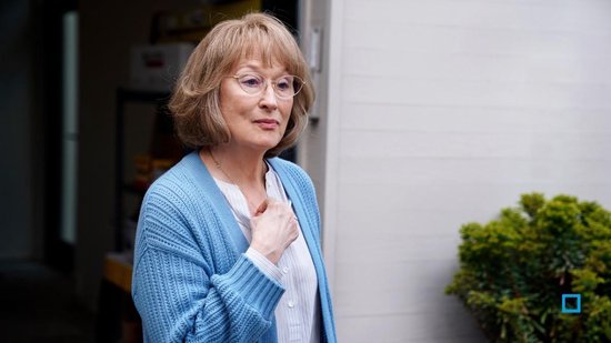 Meryl Streep met bril en een blauwe gebreide vest, staat buiten met een hand op haar borst.