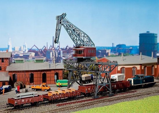 Grijze modelbouw havenkraan met rode cabine, op spoor, met wagons en vrachtwagens.