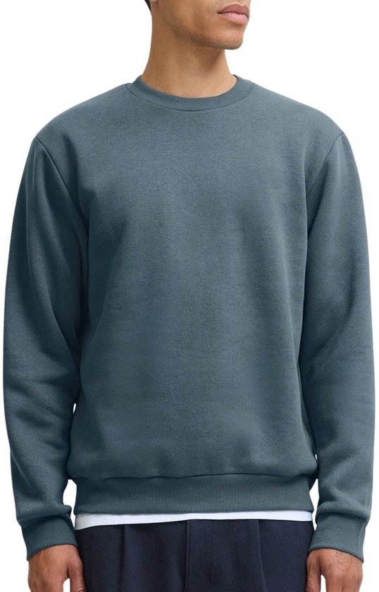 Model draagt een effen blauwgrijze sweater met ronde hals en lange mouwen. Een wit T-shirt is zichtbaar eronder.