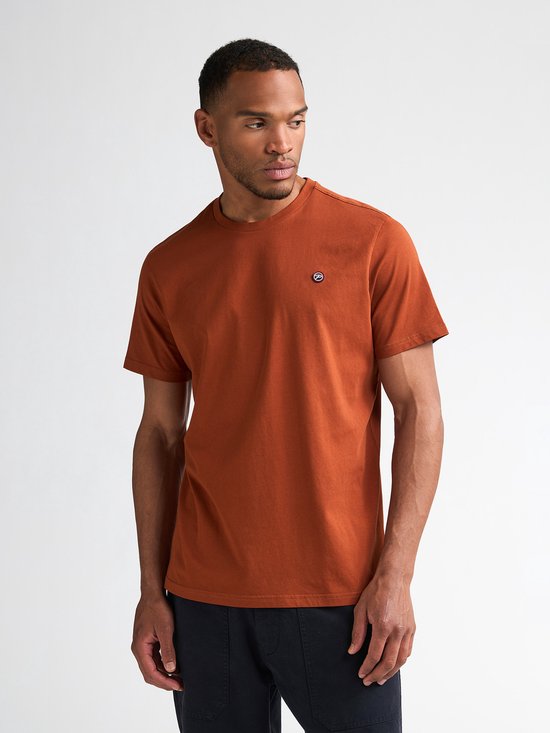 Model in een oranje t-shirt met korte mouwen en een klein rond logo op de linkerborst. Navy broek.