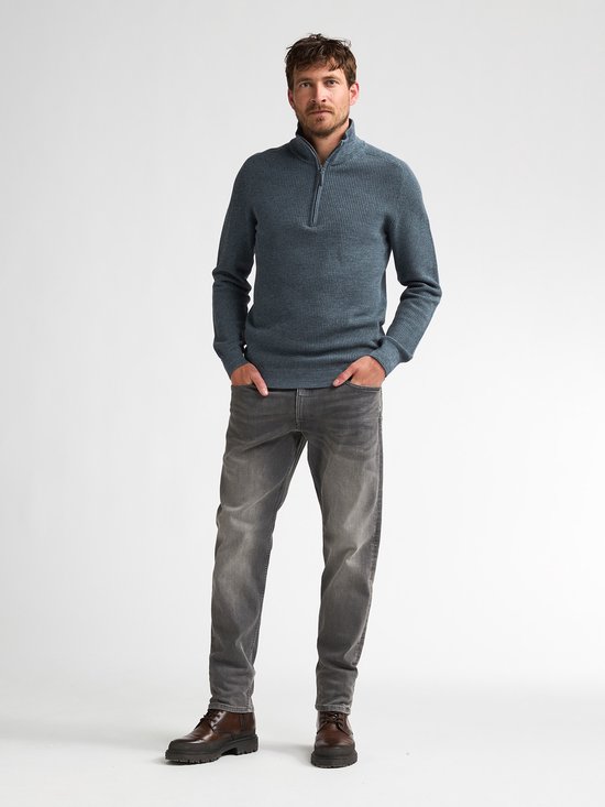 Man met grijze jeans en bruine schoenen draagt een blauwe gebreide trui met hoge kraag en ritssluiting.