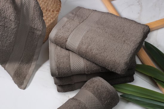 Stapel taupekleurige handdoeken met textuurranden, naast een mand en groene bladeren.