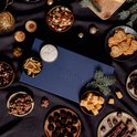 Een donkere tafel met een blauw kerstpakket en diverse borrelhapjes, kaas en chocolade.