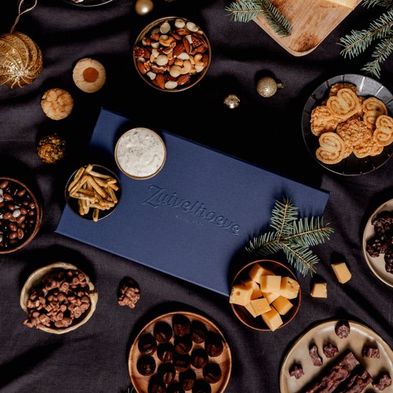 Een donkere tafel met een blauw kerstpakket en diverse borrelhapjes, kaas en chocolade.