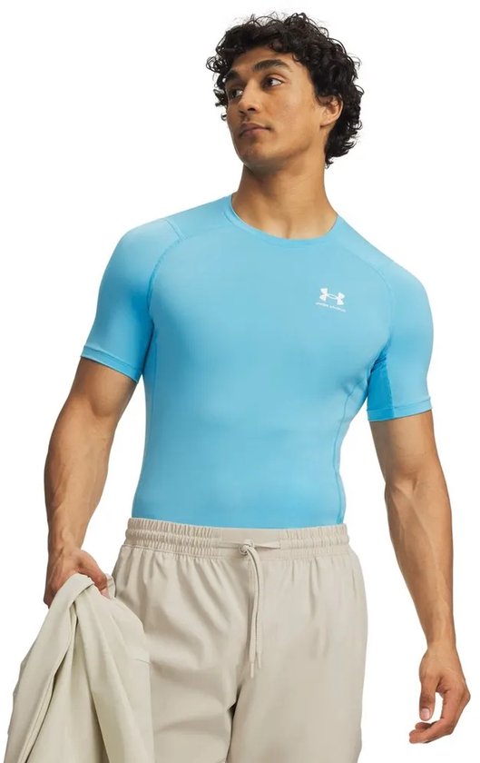 Model draagt een lichtblauwe Under Armour compressie shirt met korte mouwen en beige shorts. Met logo op de borst.