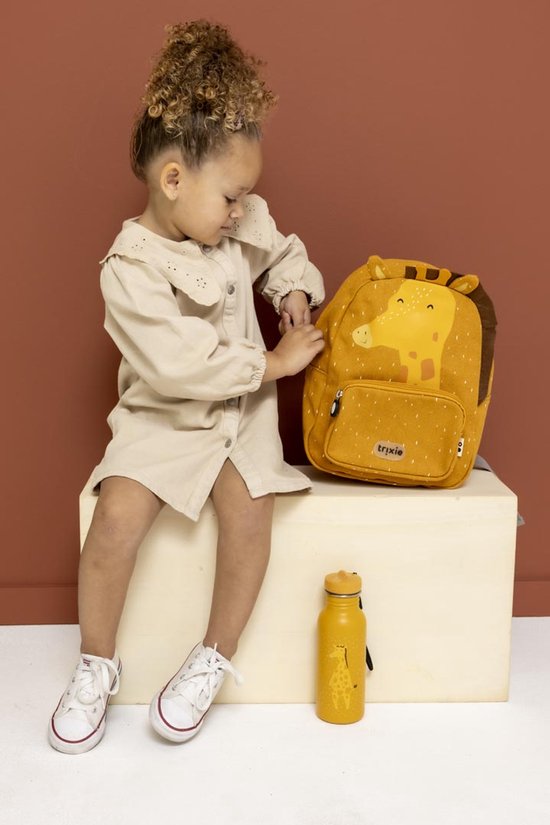 Meisje met een beige jurk zittend naast een gele 'Mr. Giraffe' rugzak met een waterfles.