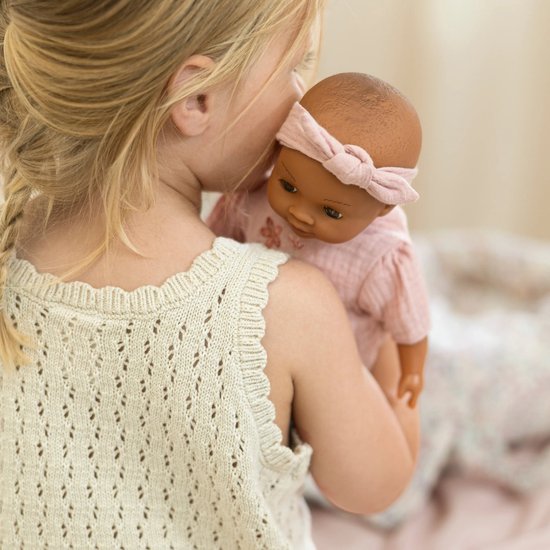 Meisje knuffelt een babypop Yara in roze outfit met bloemenborduursel.