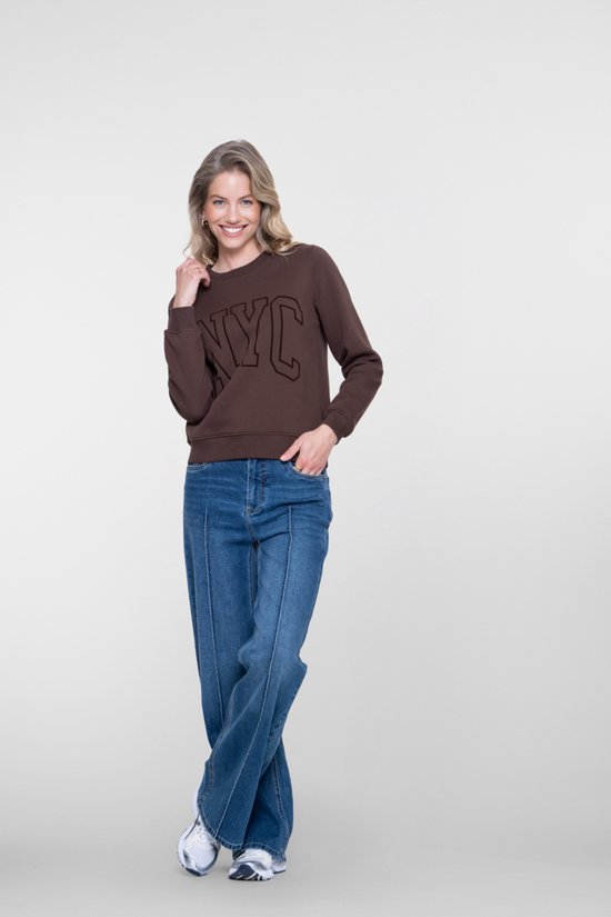 Model lacht, draagt een bruine sweater met 'NYC' opdruk en een wijde blauwe jeans. Haar ogen kijken naar de camera.