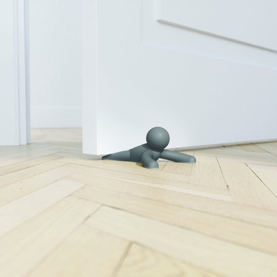 Grijze deurstopper in de vorm van een poppetje, op een houten vloer bij een witte deur.