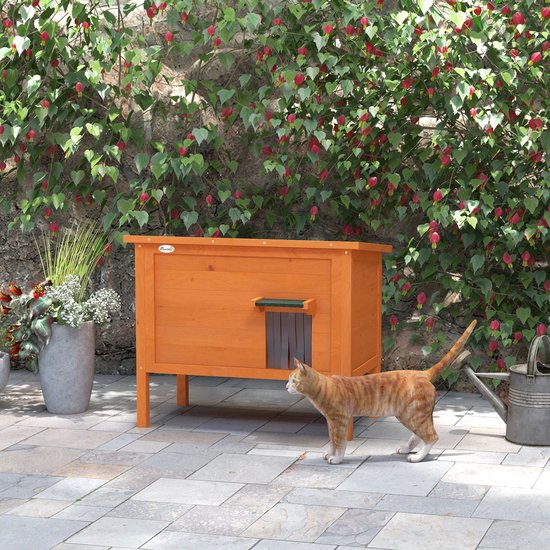 Oranje houten kattenhok in een tuin met een kat op de voorgrond en een groene struik op de achtergrond.
