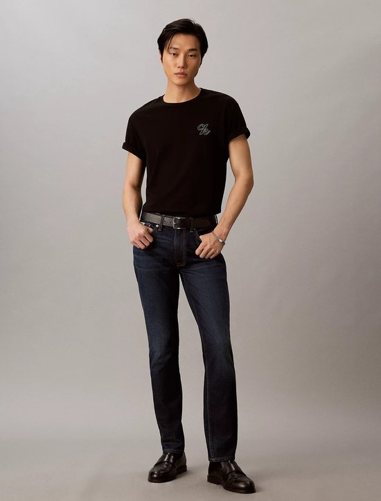 Model draagt een zwart T-shirt met CK-logo en donkerblauwe slim fit jeans met riem en zwarte instappers.