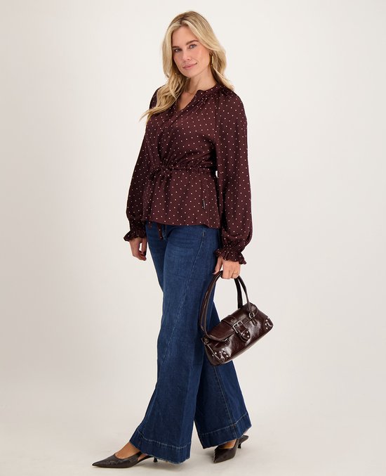 Model draagt een donkerbruine blouse met stippen, wide-leg jeans en een bijpassende handtas.