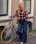 Vrouw met blond haar draagt een roze en bordeauxrood ruitjesvest en jeans bij een stadsfiets.