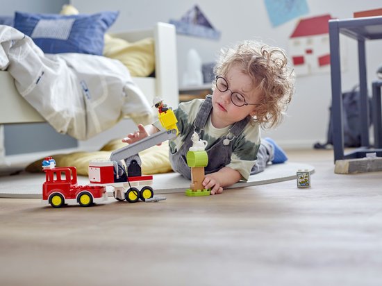 Kind met bril speelt op de grond met een rode Duplo brandweerauto, een boom en een Duplo brandweerman.