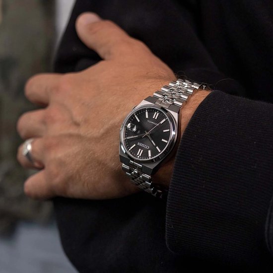 Close-up van een zilveren Citizen Tsuyosa horloge met zwarte wijzerplaat rond een pols.