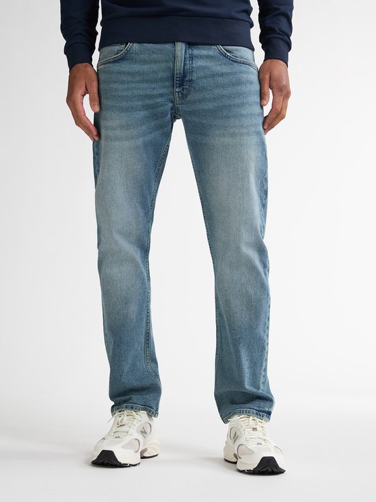 Model van voren, draagt een lichtblauwe spijkerbroek met rechte pijpen en vintage wassing. De broek is gecombineerd met witte sneakers.