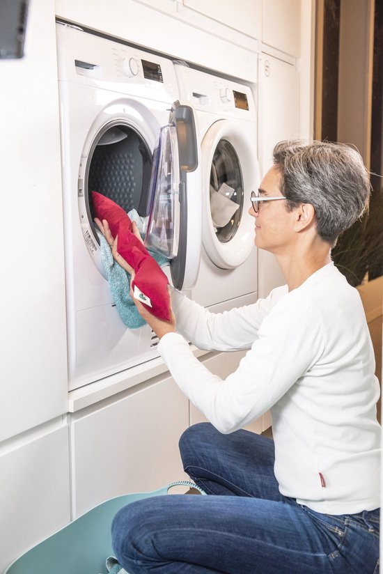 Vrouw met bril plaatst een rood kersenpitkussen in een wasmachine.