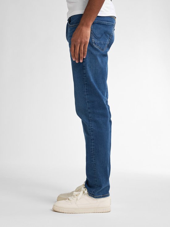 Model in een donkerblauwe stretch denim jeans, met een rechte pasvorm. Gedragen met witte sneakers.