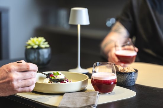 Glas met een dieprode drankje en schuimlaag op een tafel met een dessertbord en een persoon in de achtergrond.