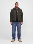 Model draagt een donkergroene jas over een bruine poloshirt, blauwe jeans en beige schoenen.