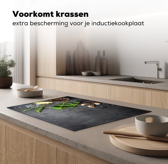 Zwarte inductie beschermer met betonprint, basilicum, rozemarijn, citroenpartjes, knoflook en peperkorrels, op een keukenblad met houten kasten.