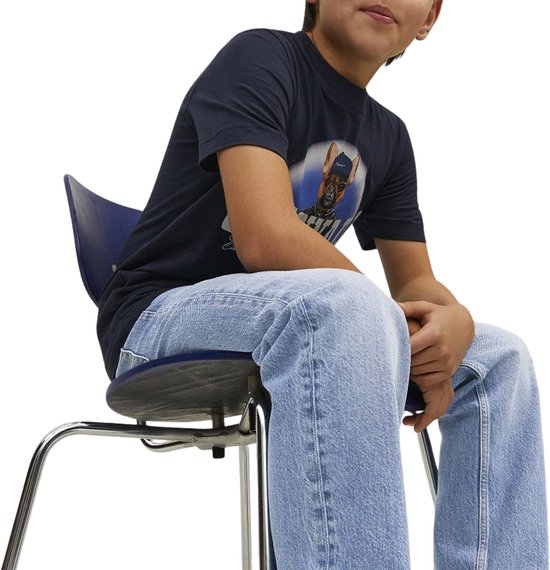 Jongen in donkerblauw T-shirt met hond-print, zittend op stoel in lichtblauwe jeans.
