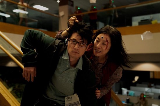 Michelle Yeoh met derde oog, houdt man met bril vast. Kantoorinterieur met trappen.