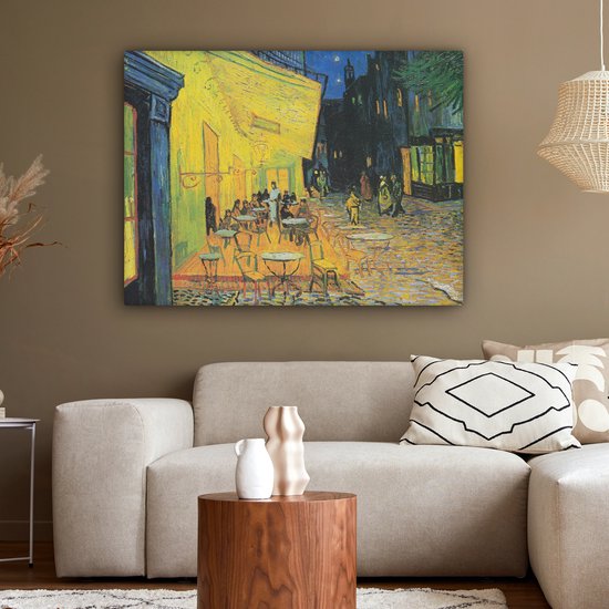 Een zithoek met een schilderij van een caféterras 's avonds, met een gele muur en tafels.