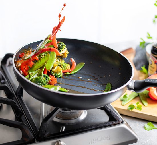 Zwarte wokpan op gasfornuis met spattende groenten en rode paprika. Handvat met "GREENPAN" logo.