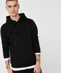 Man met tatoeages op zijn armen, draagt een zwarte hoodie met capuchon en steekzak, met witte longsleeve eronder.