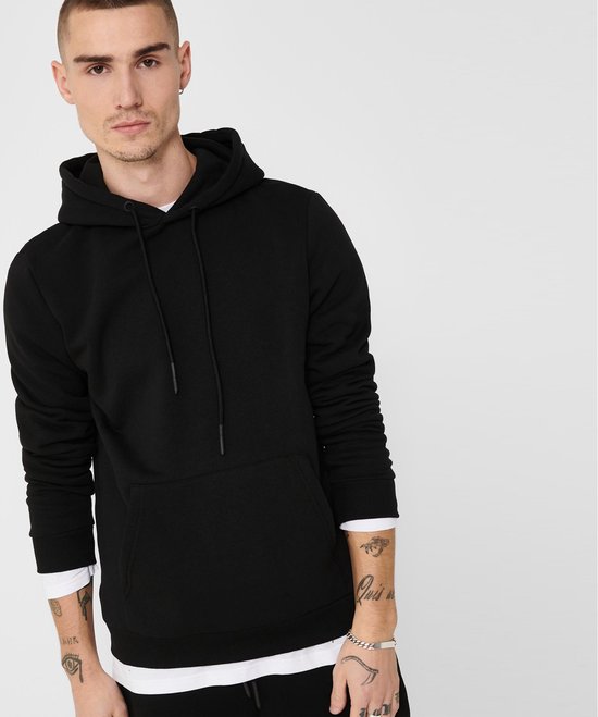 Man met tatoeages op zijn armen, draagt een zwarte hoodie met capuchon en steekzak, met witte longsleeve eronder.