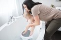 Een baby zit in een grijs badzitje in een ligbad, terwijl een vrouw met bruine top hem vasthoudt.