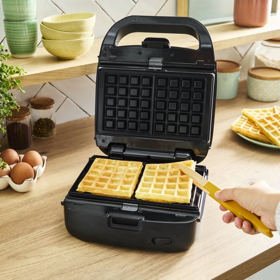 Een hand houdt een gele spatel om de gouden wafels uit een open zwart Tefal wafelijzer te halen.