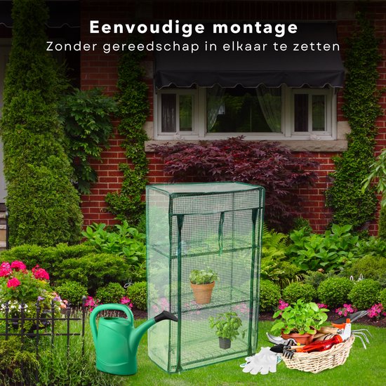 Groene kweekkas met transparante hoes, drie planken, met planten, gieter en tuingereedschap op een gazon voor een bakstenen huis.