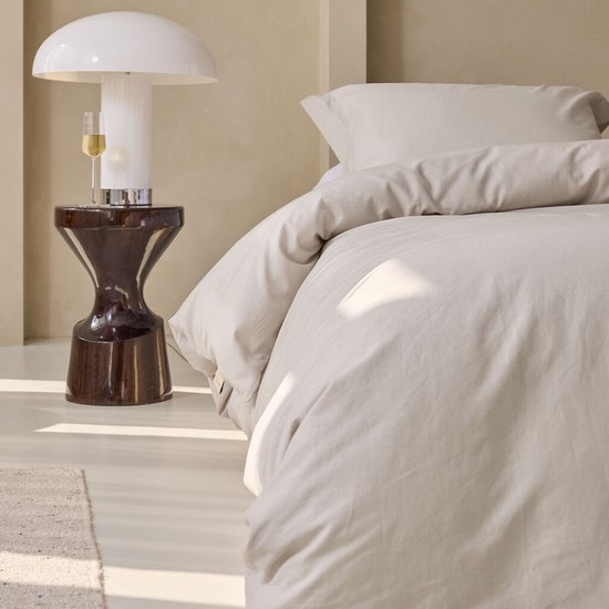 Close-up van een opgemaakt bed met zandkleurig dekbedovertrek en kussens, naast een nachtkastje met lamp.