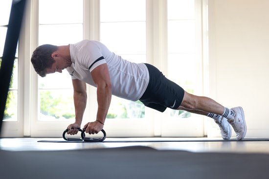 Een man in een wit shirt en zwarte korte broek doet een push-up met zwarte opdruksteunen op een mat, bij een raam.