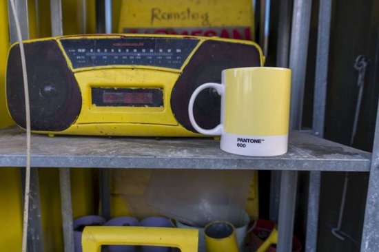 Gele mok met witte onderkant, naast een gele radio op een metalen plank.
