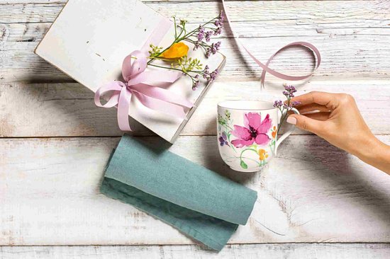Witte mok met roze bloemenprint, die door een hand wordt versierd met bloemen. Naast een cadeau.