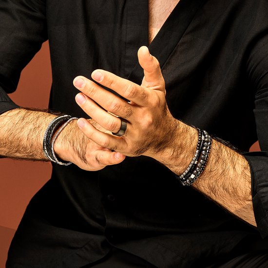 Een mannenpols met een ring en twee stapelbare armbanden. Eén armband heeft zwarte kralen, de ander een zwart gevlochten band.