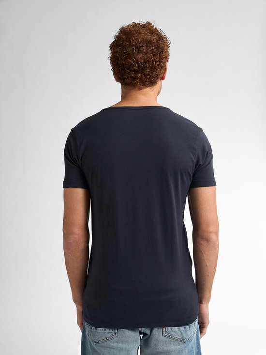 Model met krullend rood haar, sproeten, draagt een donkerblauw t-shirt en lichte spijkerbroek.