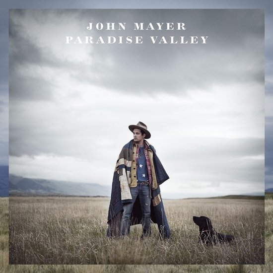 John Mayer draagt een hoed en een deken met sterrenprint. Hij staat in een veld met een hond.