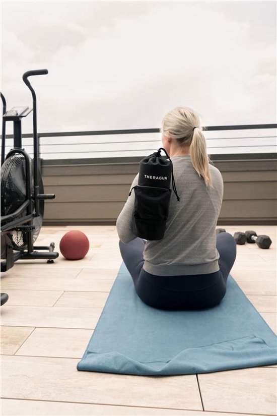 Een vrouw met blond haar zit op een blauwe yogamat op een balkon, met de rug naar de camera. Ze draagt de zwarte Theragun draagtas over haar schouder.