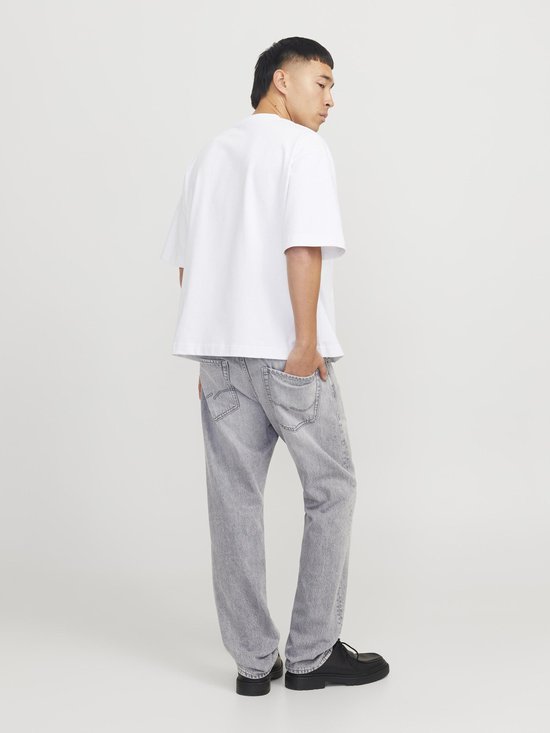 Man met korte, zwarte mullet, draagt een witte oversized top en lichtgrijze jeans. Zwart schoeisel.