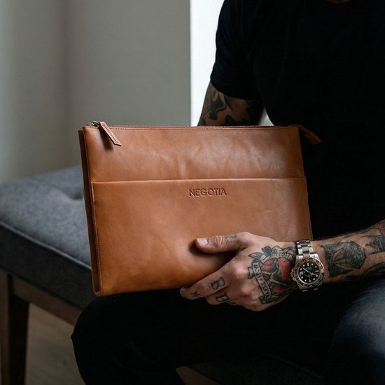 Man met tatoeages en horloge houdt een cognac leren laptoptas met 'NEGOTIA' logo vast.