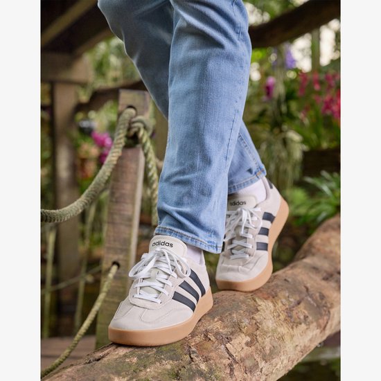 Man loopt op beige Adidas sneakers met zwarte strepen, witte veters en gumkleurige zolen, in een natuurlijke omgeving.